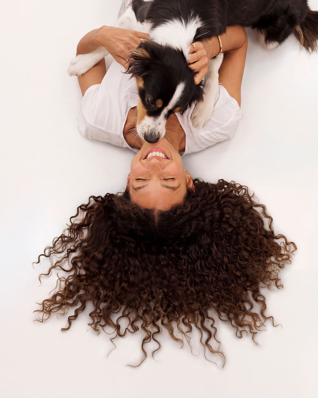 A woman lying on her back, laughing, as a tricolour dog plays on top of her